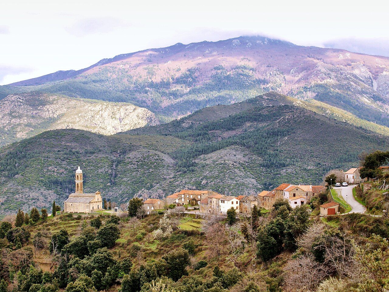 Le village d’Aiti vu depuis la route D239 : l’église Saint-Étienne et son haut clocher à gauche, les maisons alignées sur l’arête, et le Monte San Petrone en arrière-plan