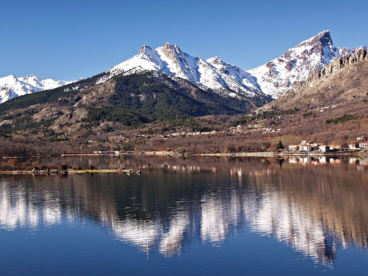 Le village d’Albertacce au pied de la Paglia Orba enneigée, vu depuis le lac de Calacuccia où se reflètent les montagnes