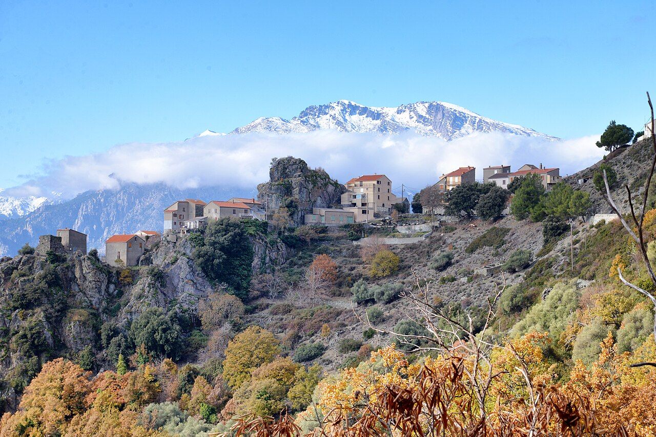 Le village d’Altiani accroché à une arête rocheuse, au milieu des arbres d’automne, avec le massif enneigé du Monte Cardo en arrière-plan