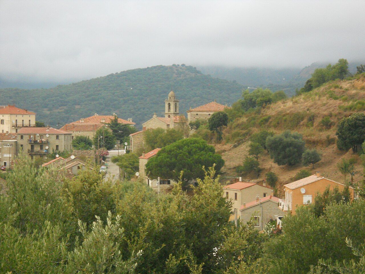 Le village d’Arbellara et le clocher de son église, à flanc de colline, sous un ciel nuageux