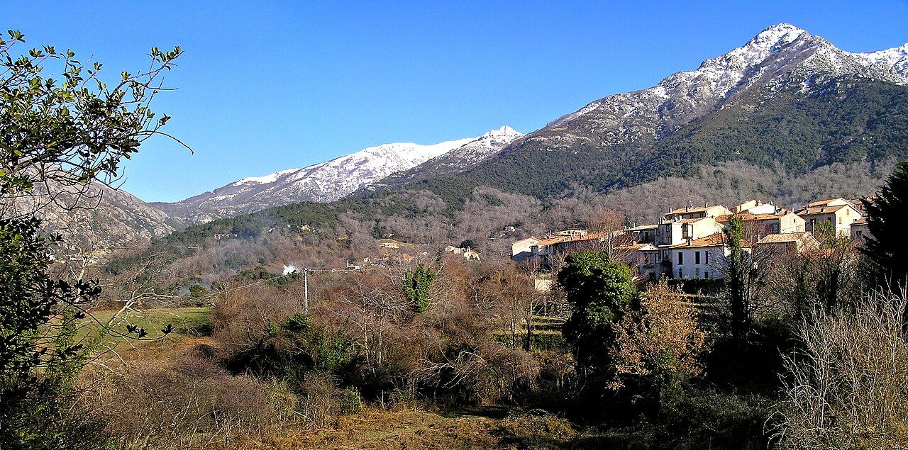 Panorama de Bocognano en hiver : maisons du village à flanc de vallée, arbres sans feuilles et montagnes enneigées menant au col de Vizzavona