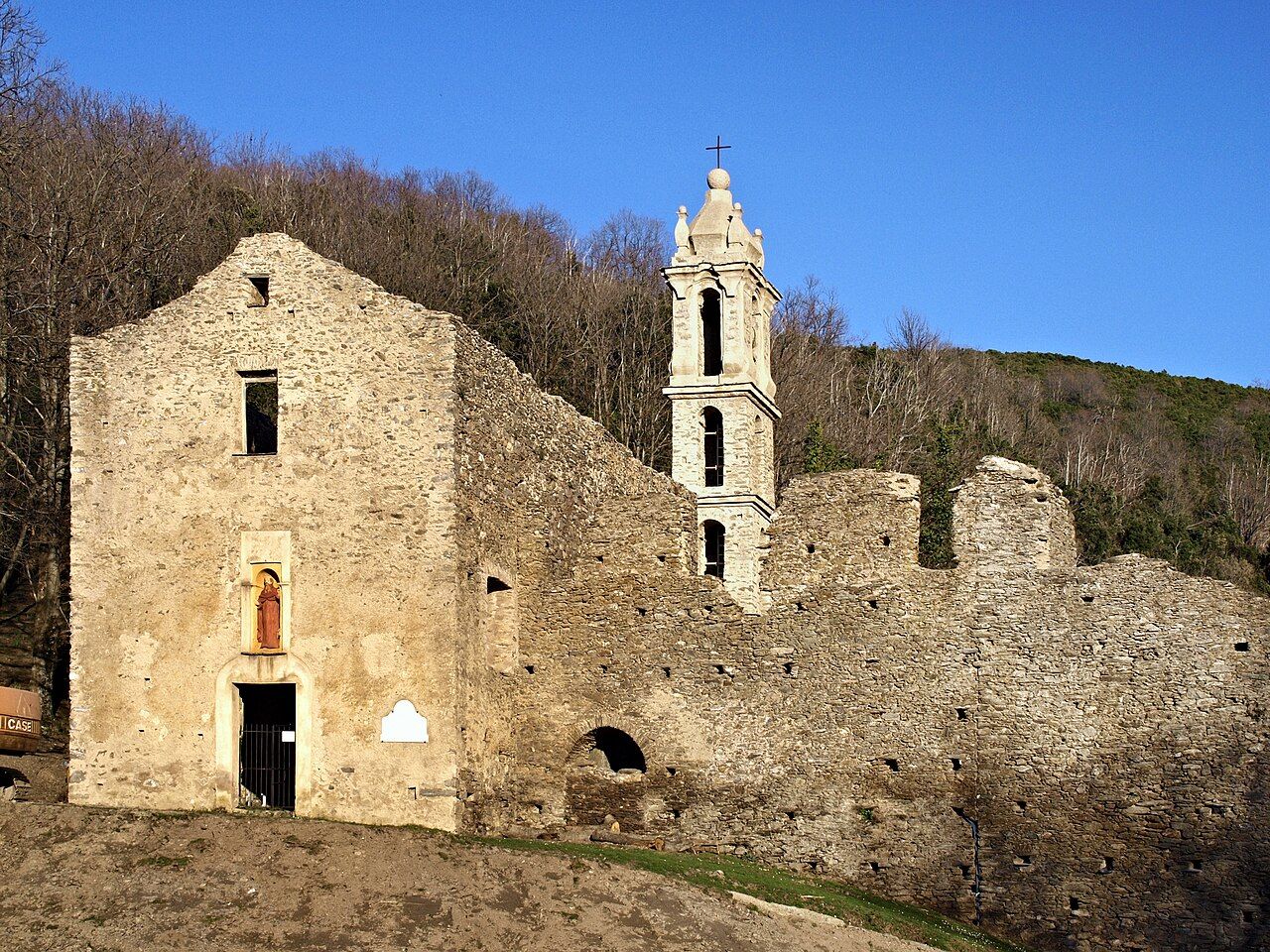 Les ruines du couvent Saint-Antoine de Casabianca, sa façade en pierre avec une statuette dans une niche et son clocher blanc, devant une forêt de châtaigniers