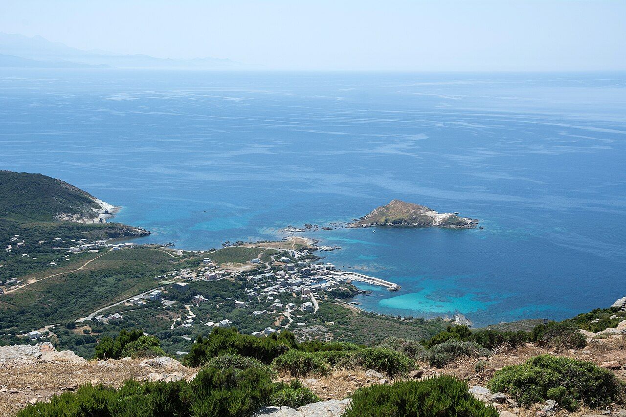 La baie de Centuri vue des hauteurs : le port et ses maisons au bord de l’eau turquoise, l’îlot de Capense au large et la mer à perte de vue