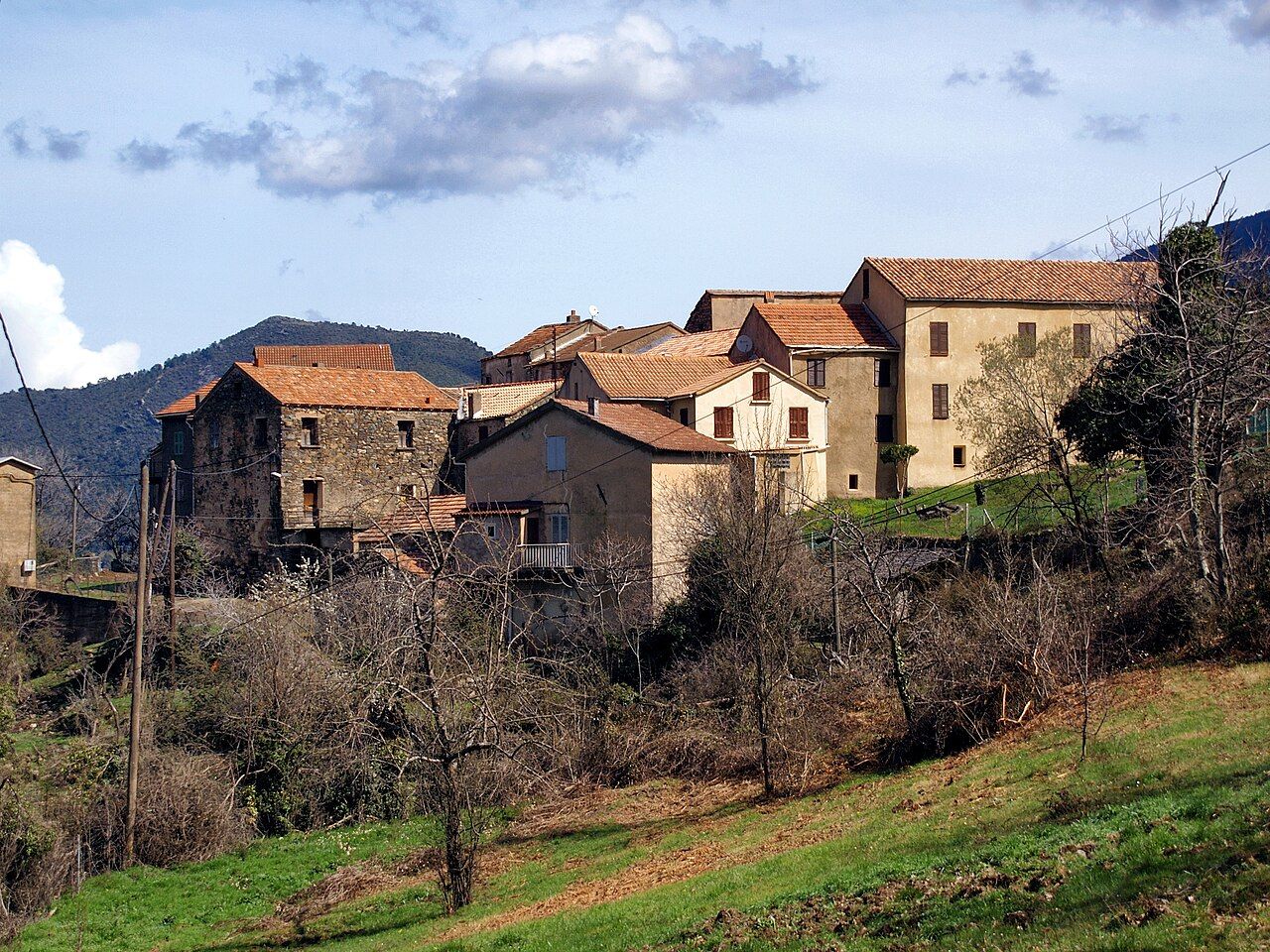 Le village d’Érone vu depuis la route D239 qui mène à Rusio : maisons de pierre et façades crépies aux toits de tuiles, au pied d’une colline boisée