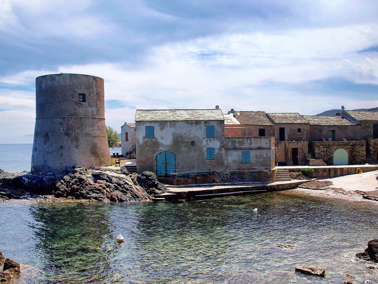 La marine de Tollare, à Ersa : une tour génoise ronde et des maisons de pierre aux toits de lauzes au bord d’une eau calme et transparente