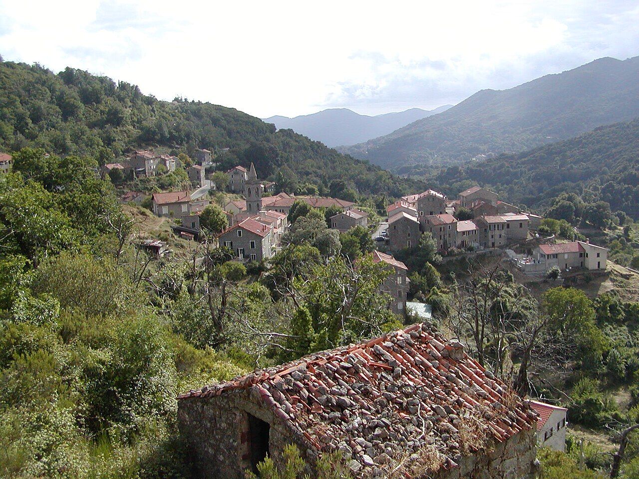 Le village de Frasseto, ses maisons de pierre aux toits de tuiles et son clocher, dans un vallon boisé entouré de montagnes ; au premier plan, une vieille cabane au toit de lauzes