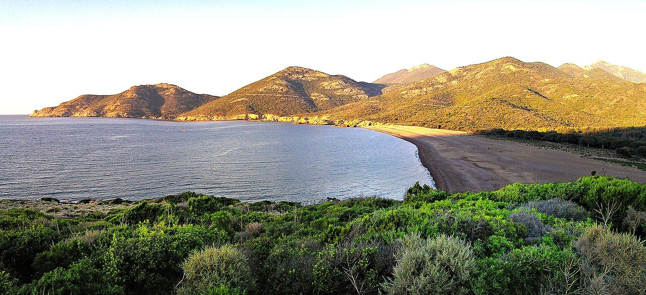 Le golfe de Galéria au soleil couchant : grande plage, eau calme et collines rouges couvertes de maquis
