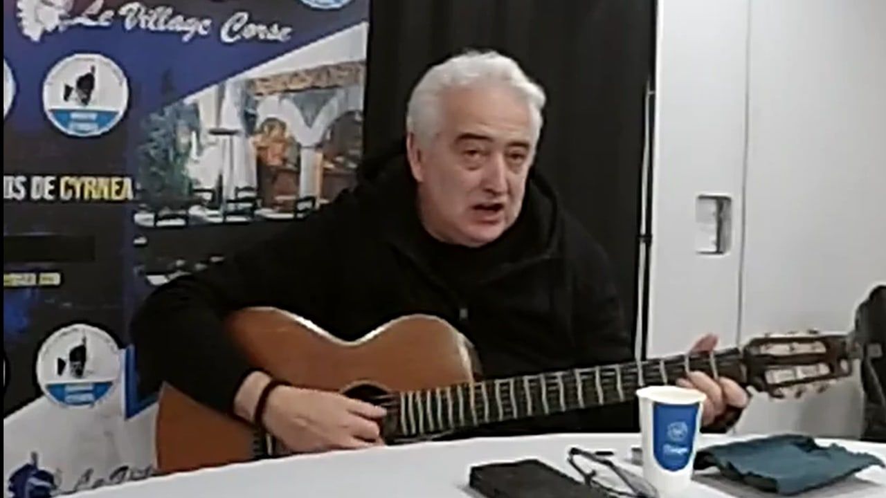 Tony Parenti, guitare à la main, chante assis à une table devant une banderole du Village Corse