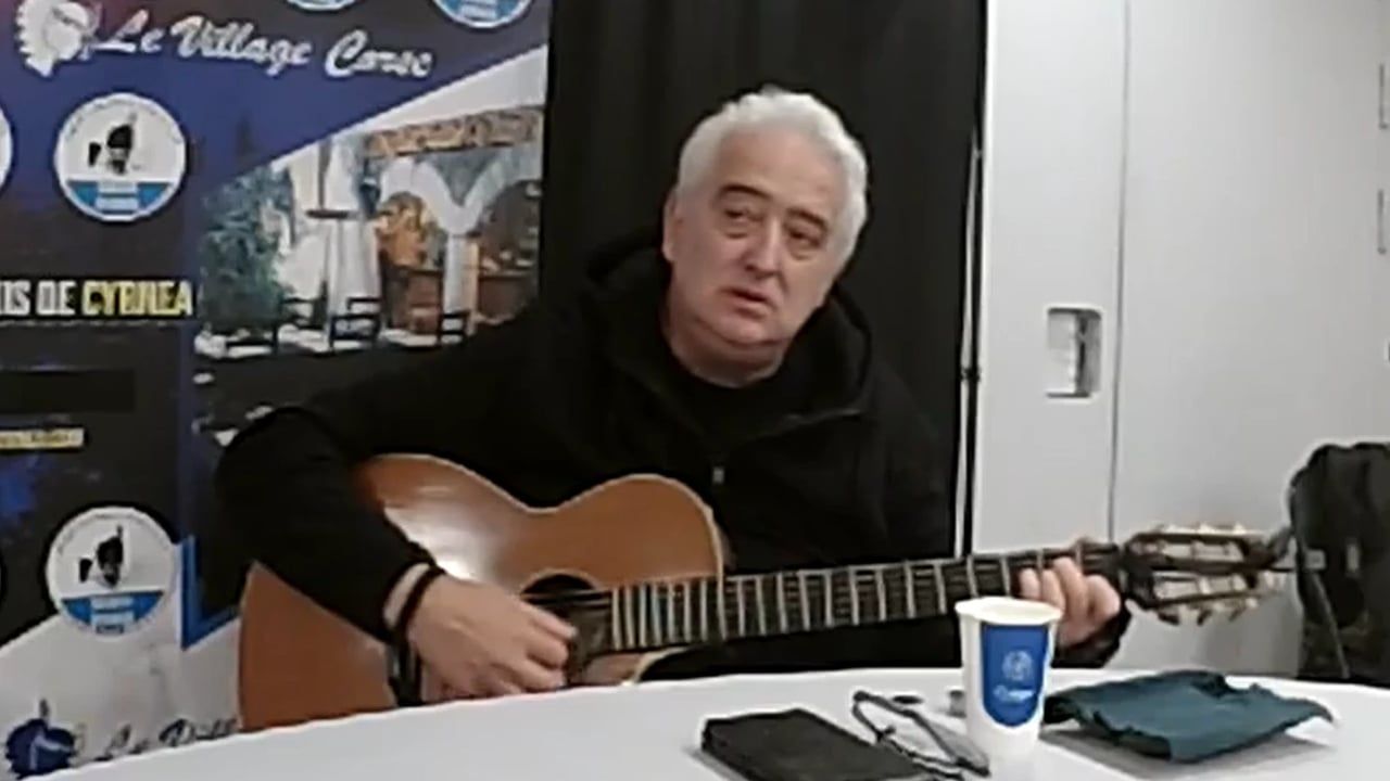 Un chanteur aux cheveux blancs joue de la guitare assis à une table, devant une banderole du Village Corse