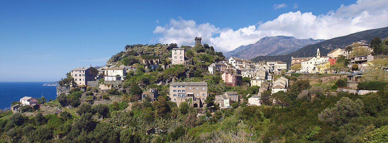 Panorama de Nonza : maisons de pierre accrochées à la pente, tour carrée au sommet, clocher, mer à gauche et montagnes du cap Corse au fond