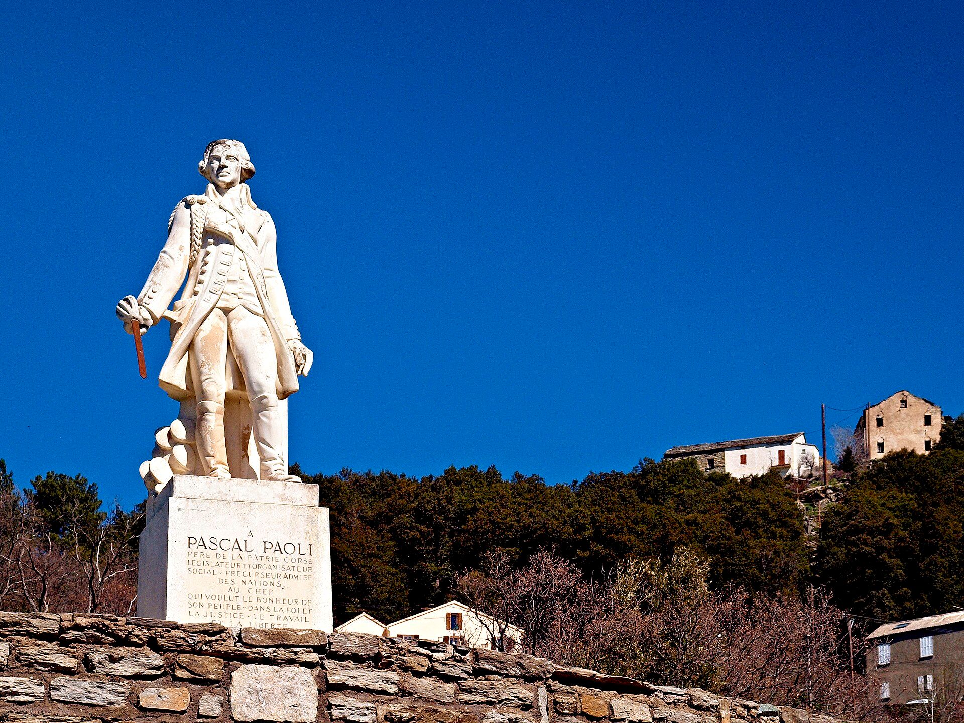 Statue blanche de Pascal Paoli sur son piédestal, devant le ciel bleu et les maisons de Morosaglia, son village natal