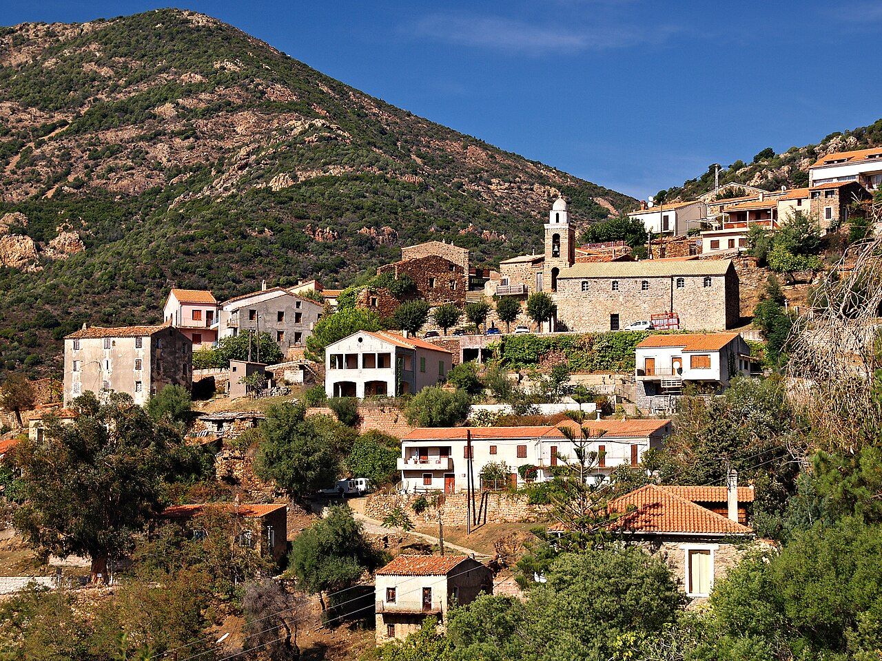 Le village de Partinello étagé sur une colline, avec son église à clocher, au pied d’une montagne de maquis et de rochers rouges