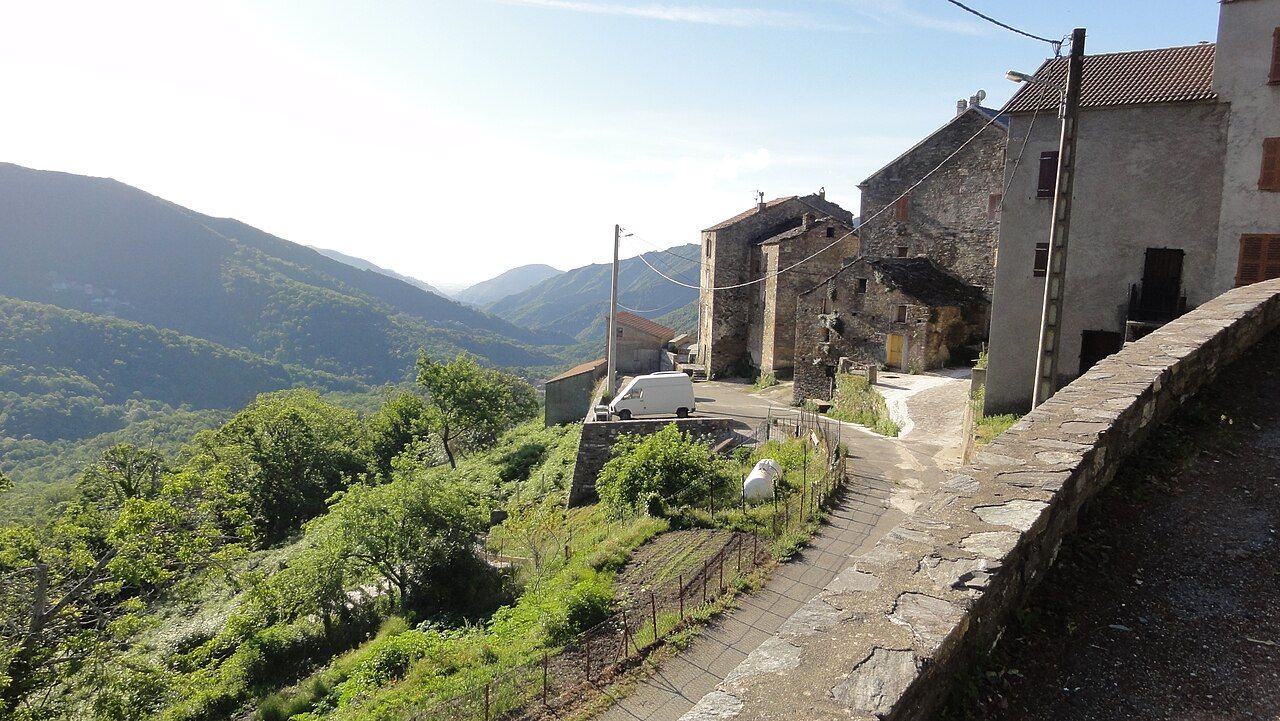 Maisons de pierre d’un hameau de Perelli au bord d’une ruelle en pente, face aux montagnes boisées de la vallée de l’Alesani
