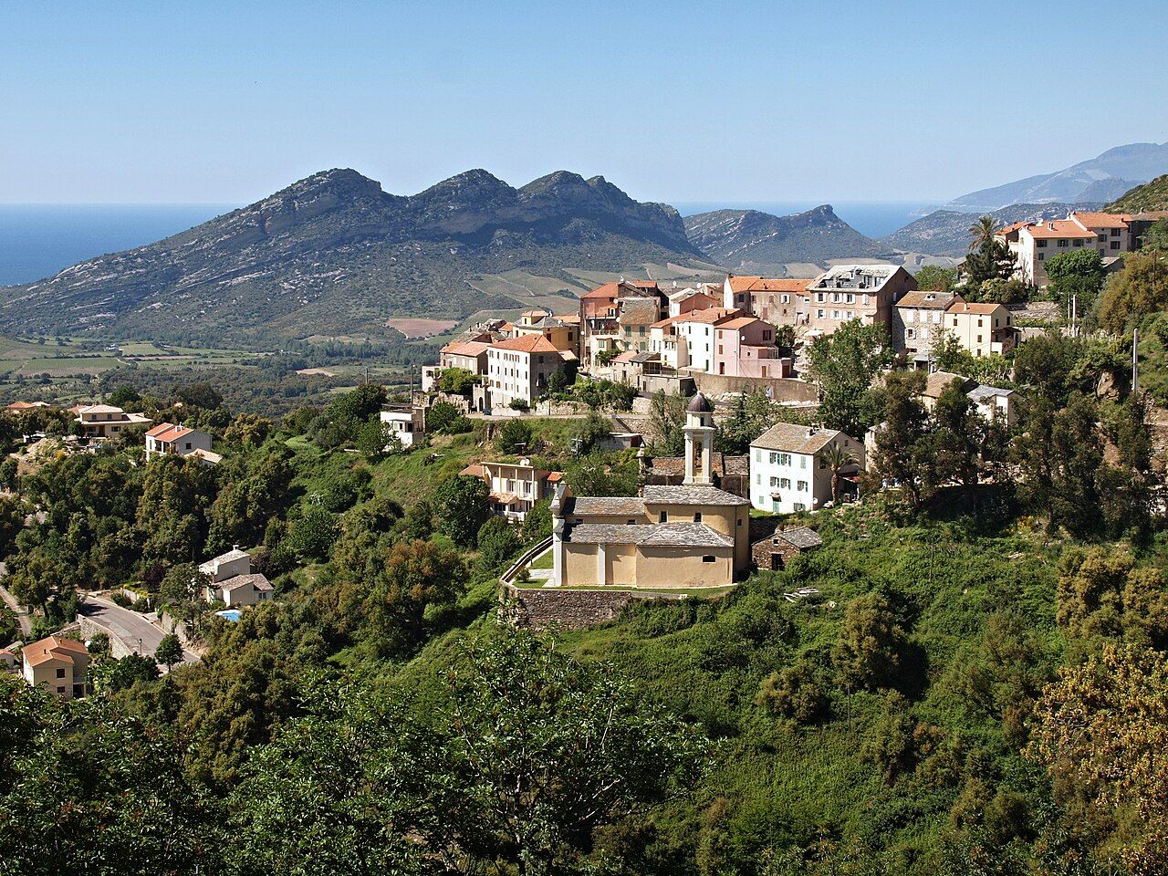 Le village de Poggio-d’Oletta et son église San Cervone sur un versant boisé, avec la plaine du Nebbiu, les collines calcaires et la mer au fond