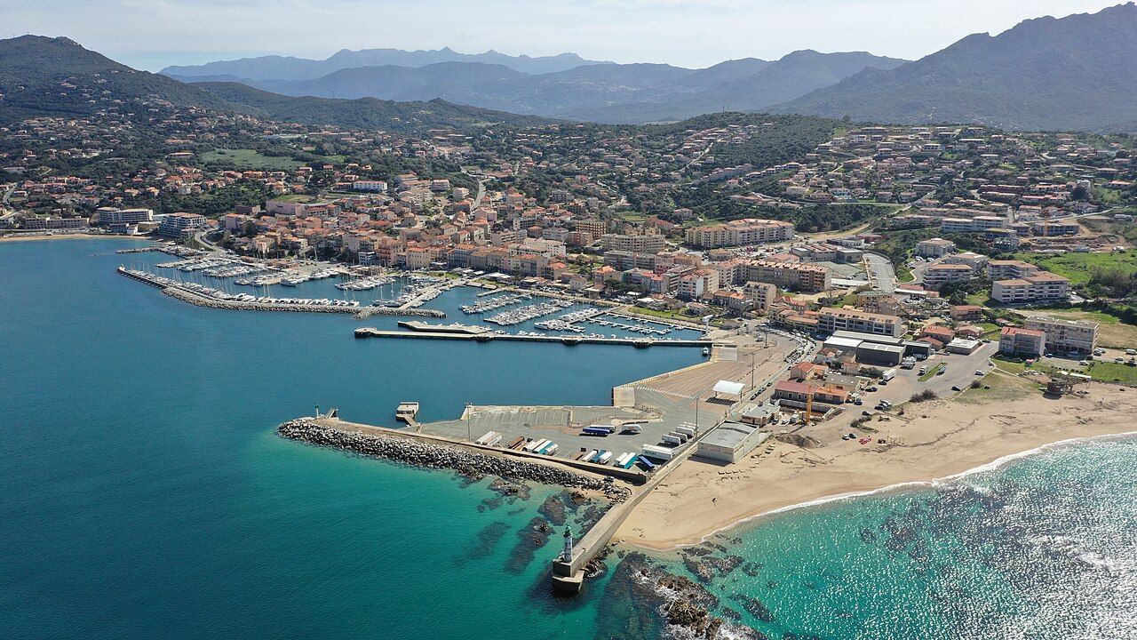 Vue aérienne de Propriano : la ville, le port de commerce avec sa jetée et son phare, le port de plaisance et la plage, montagnes en arrière-plan