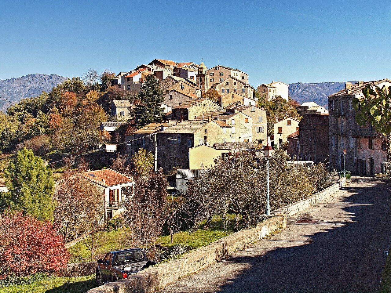Le village de Riventosa, ses maisons serrées sur un éperon autour du clocher, vu depuis la route sous un ciel bleu