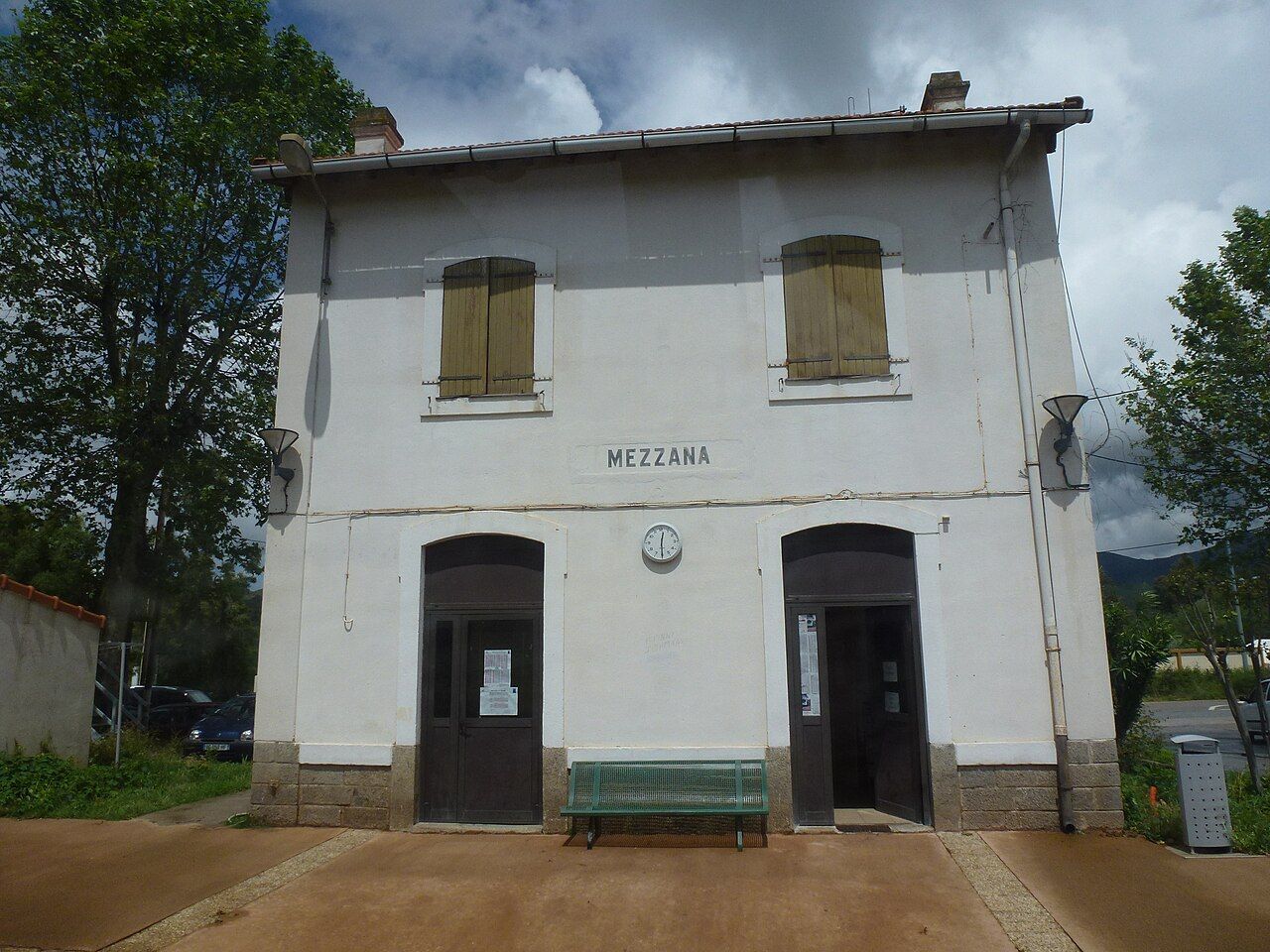 La gare de Mezzana, sur la commune de Sarrola-Carcopino : bâtiment blanc à un étage, volets en bois, horloge et banc vert, sous un ciel nuageux