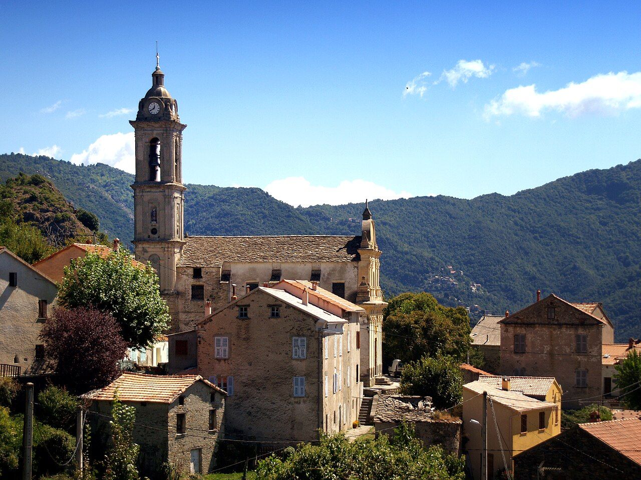 Le village de Sermano et l’église paroissiale de l’Annonciation, son haut clocher et son toit de lauzes, devant les montagnes boisées du Boziu