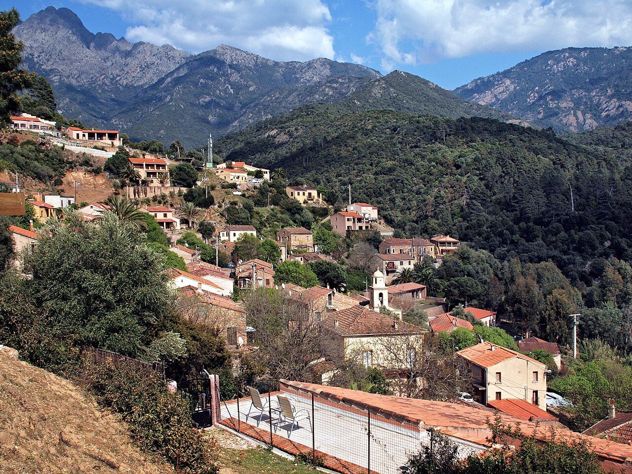 Les maisons et le clocher de Serriera étagés sur un versant boisé, au pied de montagnes rocheuses