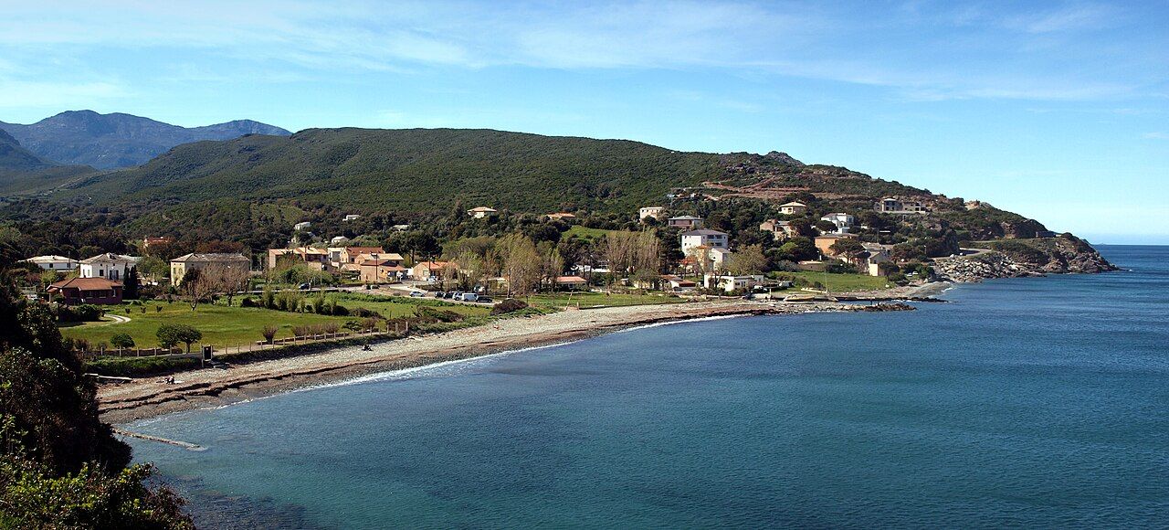 La marine de Sisco : une plage de galets en arc de cercle, des maisons dans la verdure et les collines du cap Corse au-dessus de la mer