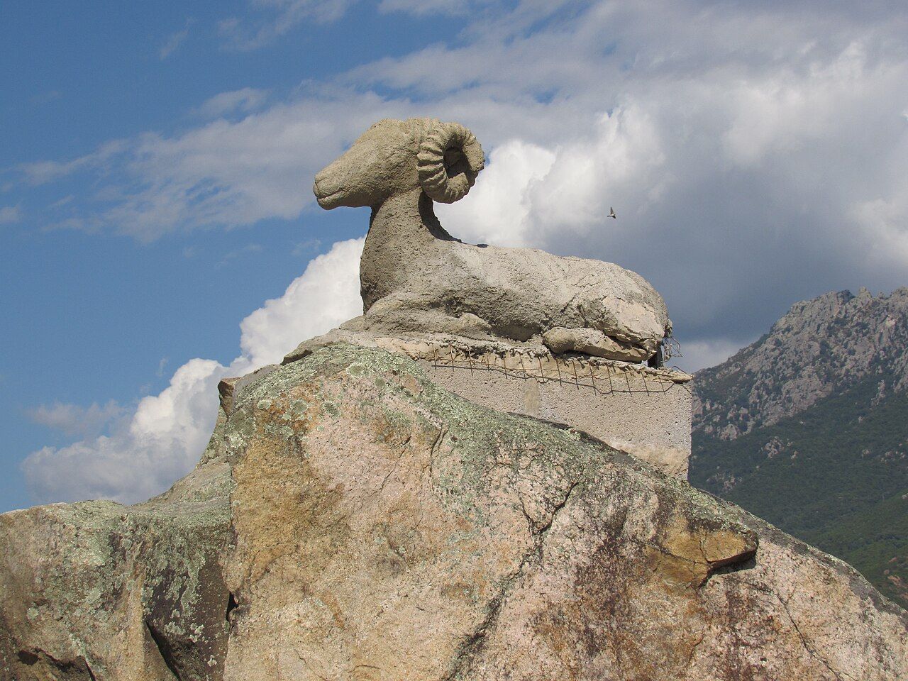 La sculpture « U muvrinu », un mouflon couché posé sur un rocher de granit à Tavaco, devant des montagnes couvertes de maquis et un ciel nuageux